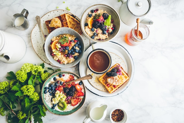 A selection of breakfast options including a smoothie bowl topped with fruits, a couple of fruit-topped oat bowls, a few slices of toast, a cup of tea with honey and milk on the side.