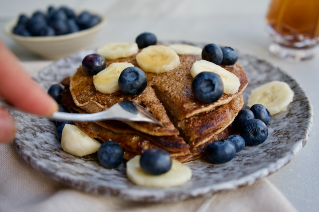 A hand holding a fork, sticking into a plate of banana pancakes garnished with blueberries and banana slices