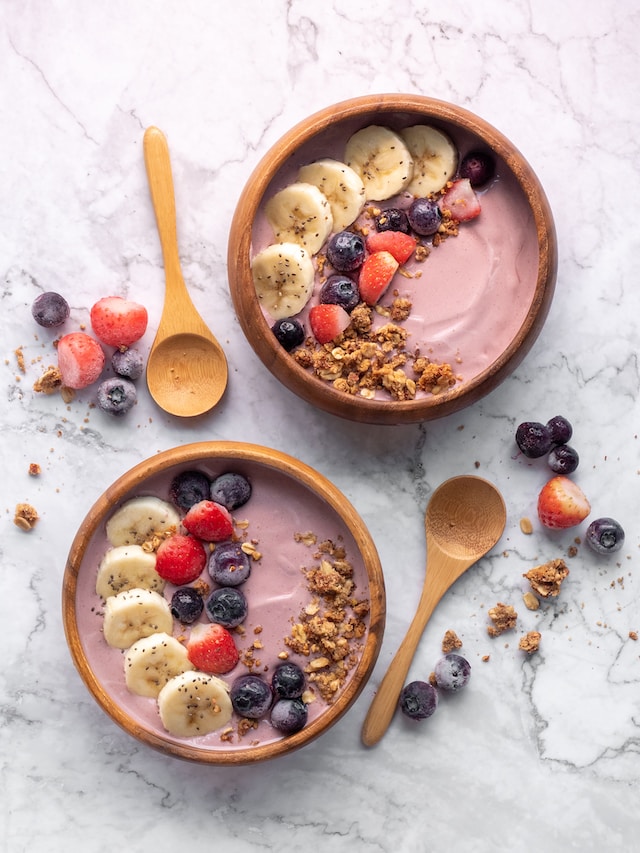 A couple of wooden smoothie bowls topped with bananas, strawberries, blueberries and nuts, with two wooden spoons on the side