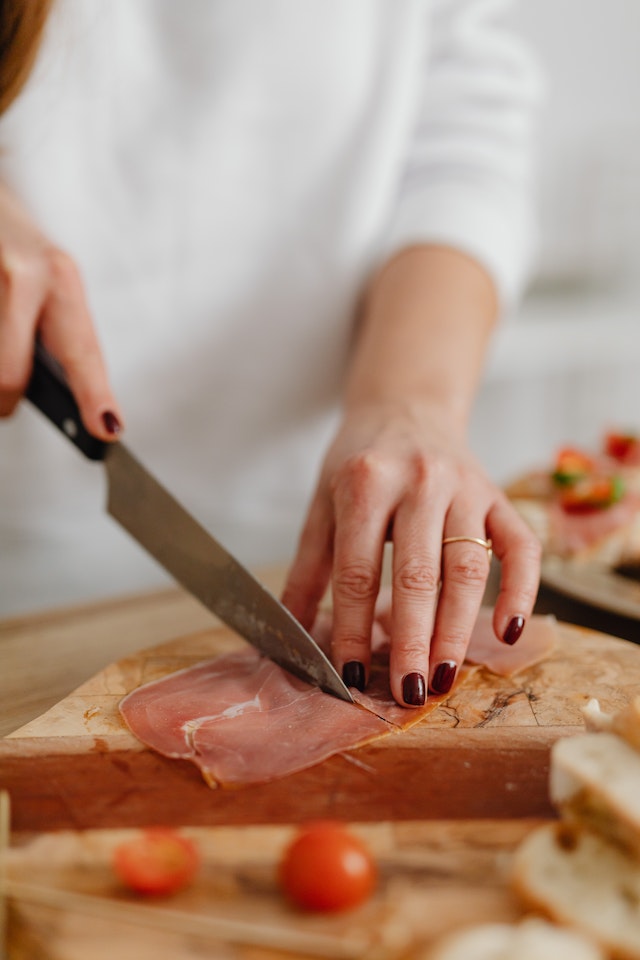 A person slicing a thin piece of ham on a wooden board